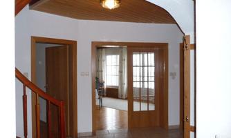 A bright entrance area with a staircase and wooden details. In the background are large windows that offer a view of another room.