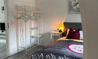 A cozy bedroom with a bed, a nightstand, and a shelf. The walls are light and the floor is wooden.