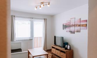 A cozy living area with a dining table, chairs, and a television. Large windows provide plenty of natural light.