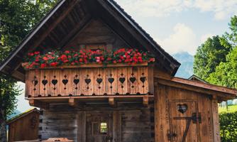 A charming wooden house with a balcony full of flowers. It is situated in a green environment under a blue sky.
