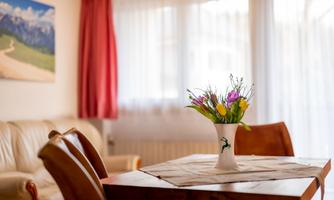 A cozy room with a wooden table and colorful flowers in a vase. In the background, there is a bright window and a picture of mountains.