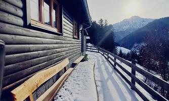 A cozy cabin in the mountains with snow and a wooden bench. The sun gently shines behind the mountains.