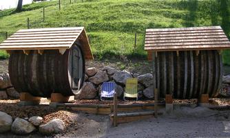 Two wooden accommodations that look like large barrels are outside. Between them are two deck chairs and in the background, a green meadow can be seen.