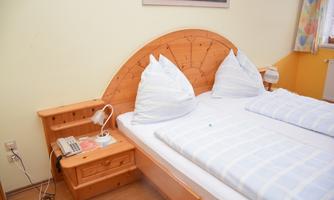 A cozy bedroom with a double bed and wooden frame. The pillows are stacked high and there is a bedside table with a lamp.