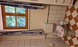 A small bathroom with a toilet and a window. The walls are tiled and there is a white cabinet.