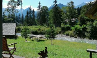 A peaceful landscape with trees and mountains in the background. A small river flows through the meadow, where a bench and a chicken decoration are located.