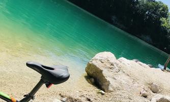 A bicycle stands by the shore of a clear, green lake. In the background, trees and a blue sky can be seen.