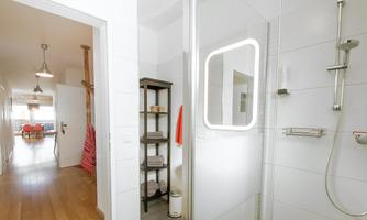 A modern bathroom with a shower and a round mirror. In the background, a stylish hallway with wooden flooring and light sources is visible.