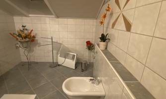 A bathroom with white tiles and a gray floor. There is a bathtub, plants, and a flower arrangement in one corner.