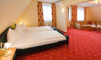 A cozy hotel room with two beds and a seating area. The carpet is red with patterns, and large windows provide plenty of daylight.