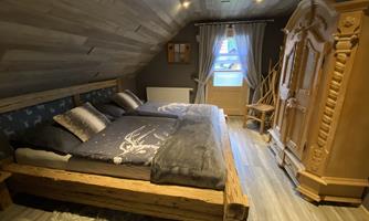 A cozy attic room with a large wooden bed and decorative pillows. The room is paneled in wood and has a rustic wardrobe as well as a window with curtains.