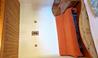 A small bedroom with a wooden bed, an orange sofa, and a wooden child’s bed. The walls are plain and there are some pictures hanging.