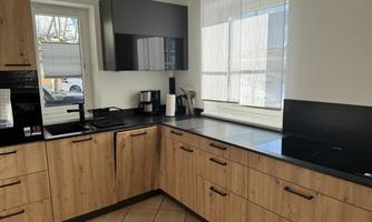 A modern kitchen with wooden cabinets and black countertops. Large windows provide plenty of light and a bright atmosphere.