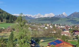 A picturesque landscape with green meadows and small houses. In the background, majestic mountains rise under a clear blue sky.