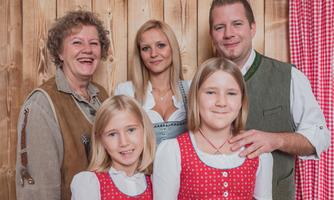 A cheerful family poses in front of a wooden wall. They are wearing traditional clothing, including dirndls and folk jackets.