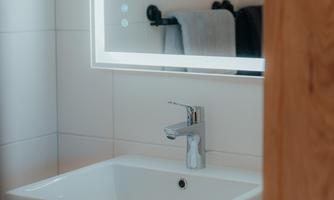 A modern bathroom with a square sink and a lit mirror. Towels are hanging on the wall in the background.
