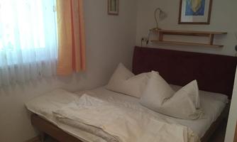 A simple bedroom with a double bed and two large pillows. The room has a window with curtains and a bright wall decoration.