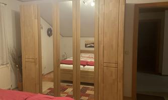 A cozy bedroom with a wooden wardrobe and a double bed in the background. The walls are light and the decoration is simple.