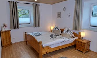 A cozy bedroom with a wooden bed, two nightstands, and soft pillows. Large windows let in plenty of daylight, and the walls are designed in light colors.