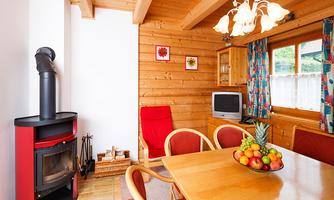 A cozy living space with wooden walls and a table decorated with fresh fruit. In the background, there is a fireplace and a television, as well as colorful curtains.