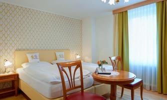 A cozy bedroom with two single beds and a round table. The walls are decorated with a modern pattern and plenty of light streams through the windows.