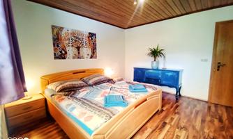 A cozy bedroom with a double bed and wooden floor. A colorful picture hangs on the wall, and a plant sits on the dresser.