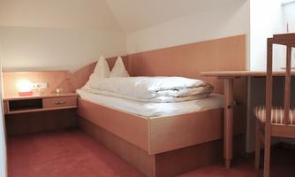 A simple room with a single bed and wooden shelves. There is a table and chair as well as a bedside lamp.