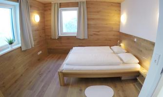 A cozy bedroom with wooden paneling and a large windowsill. The bed is simple and inviting, surrounded by soft colors.