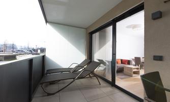 A modern balcony with two loungers and plenty of daylight. In the background are large windows that overlook a bright living space.