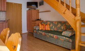 A cozy living room with a sofa and a wooden staircase. The walls are light and there is a wooden dining table.