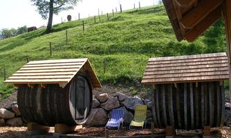 Two round wooden houses with wooden shingle roofs stand on a green meadow. In the foreground, two deck chairs are placed.