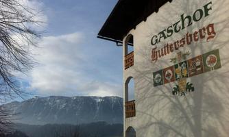 A guesthouse with the inscription "Huttersberg" and a picturesque mountain panorama in the background. The sky is partly cloudy, and the landscape looks calm and inviting.
