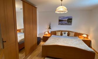 A bright bedroom with a large double bed and wooden wardrobes. A carpet and a beautiful picture on the wall are visible.