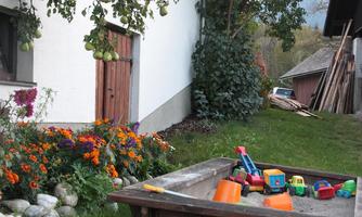 A sandbox with colorful toys stands in a garden next to a white wall and blooming flowers. In the background, trees and an outbuilding can be seen.