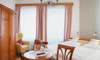 A cozy hotel room with two beds and a round table. Large windows let in plenty of light, and the decor is friendly and inviting.