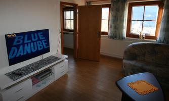 A cozy living space with a television displaying "BLUE DANUBE". Along with it, there is a couch, a side table, and windows with a view outside.