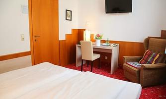 A cozy room with two beds, a desk, and a TV on the wall. A comfortable seating area with an armchair and a padded blanket is also available.