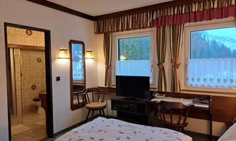 A cozy room with two beds, a television, and a desk. The room has large windows overlooking nature and an adjacent bathroom.