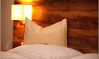 A cozy bed with pillows and a warm wooden panel in the background. A soft lamp provides atmospheric light.