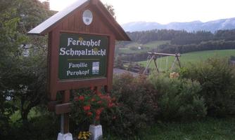 A sign of the holiday farm Schmalzbichl with the name of the Pernkopf family. In the background, gentle hills and a calm sky can be seen.