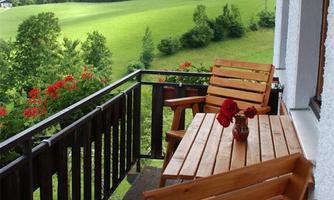 A balcony with wooden furniture and red flowers. In the background, a green landscape extends with mountains.