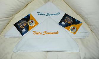 Two folded towels with the inscription "Villa Sonnwend" are lying on a pillow. Next to them are two postcards featuring images of the house.