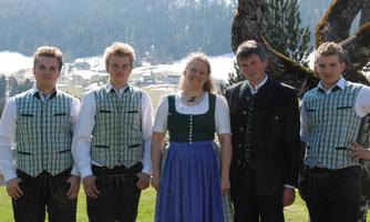 A group of four people in traditional clothing stands on a meadow. In the background, snow-capped mountains and a clear sky are visible.