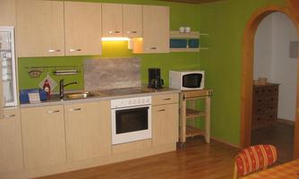 A modern kitchen with bright wooden furniture and green walls. It includes a sink, a stove, a microwave, and a dining table with a checkered tablecloth.