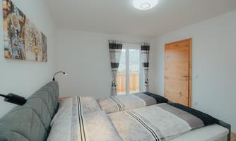 A bright bedroom with two single beds and modern furniture. Large windows provide access to a balcony and plenty of natural light.