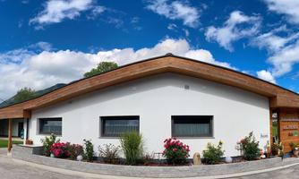 A modern house with a wooden roof and white walls. In front of the house, there are colorful flowers and a clear blue sky.