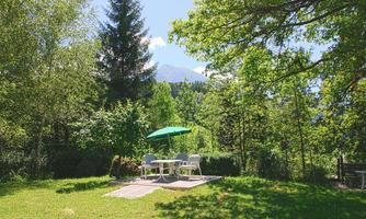 A beautiful garden with a table and chairs under a green umbrella. In the background, trees and mountains can be seen.