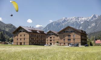 A modern building in a picturesque mountain landscape. A paragliding pilot glides over a blooming meadow.