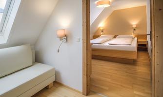 A bright bedroom with wooden finishes and soft light sources. In the background, a comfortable bed with white bedding can be seen.