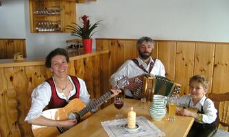 A musical group is sitting at a wooden table. The woman is playing the guitar, the man is playing the accordion, and a boy is sitting next to them while they play songs.